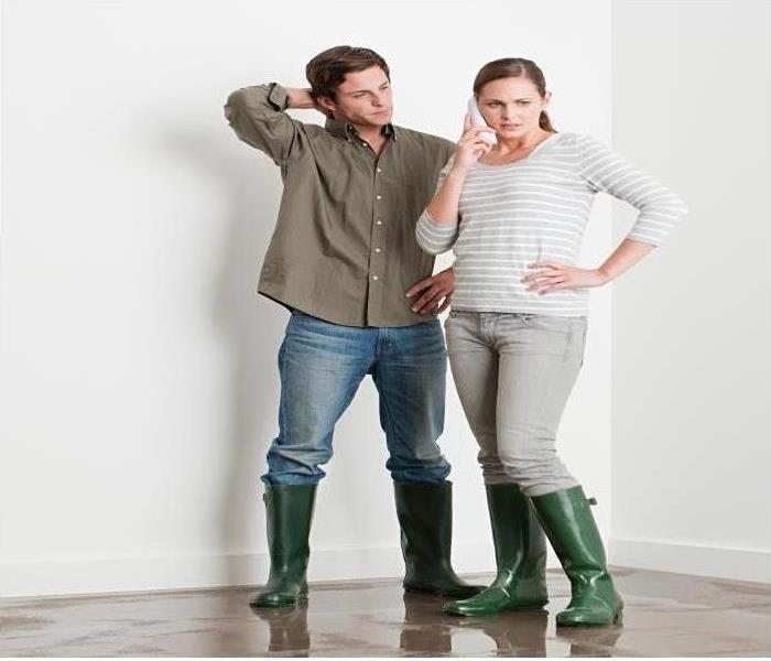 man and woman in rain boots standing in water damaged room, pondering their next step